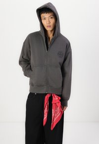 Sweat à capuche zippé gris avec un patch logo, poches avant et cordon de serrage à la capuche. Assorti à un pantalon noir et un bandana rouge noué à la taille.