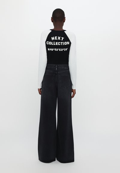 Long-sleeve black and white top featuring text "NEXT COLLECTION" on the back. Paired with wide-leg dark denim jeans.