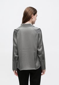 Svilena siva bluza s ovratnikom, raglan rukavima i blago produljenom stražnjom šavom, uparena s crnim hlačama.