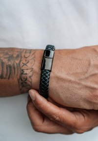 Hand with tattoo adjusting a black braided leather bracelet with a shiny black magnetic clasp on wrist.