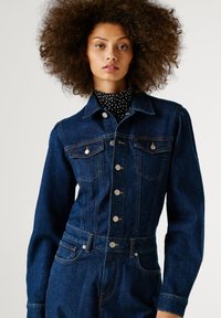 Džínová bunda v tmavom praní so striebornými gombíkmi, dvoma náprsníkovými vreckami a vystuženými ramenami, s hladkou textúrou a prispôsobeným strihom.