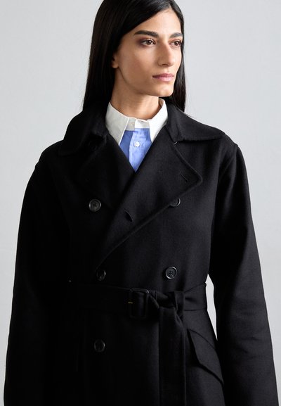 Μαύρο μάλλινο παλτό με διπλό κούμπωμα, μεγάλο γιακά και ζώνη στη μέση. Διαθέτει κουμπιά και φοριέται πάνω από ένα ανοιχτό μπλε πουκάμισο.