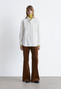 White button-up shirt with a ruffled collar, layered over a yellow turtleneck. Paired with brown corduroy flared pants. Black shoes.