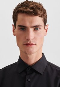 Giovane uomo con capelli castani corti e mossi e occhi azzurri, indossa una camicia nera con bottoni su uno sfondo neutro.