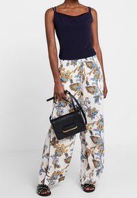 Top senza maniche blu navy con scollatura drappeggiata, abbinato a pantaloni bianchi svasati con un motivo a paisley blu e oro. Borsa nera con accenti dorati.