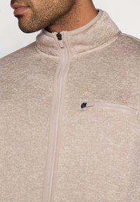 Μπεζ φλις μπουφάν με φερμουάρ, ψηλό γιακά, υφή υφάσματος και μικρή τσέπη με φερμουάρ. Διαθέτει διακριτικ ό μοτίβο με ανάμεικτο χρώμα.