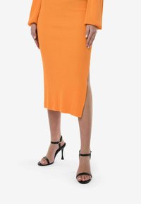 Vestido naranja con canalé y una abertura lateral, combinado con tacones negros de tiras. El vestido presenta mangas largas y una silueta ajustada.