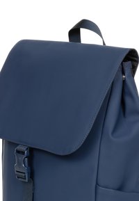 Mochila azul marino con solapa plegable, correa ajustable y cierre de hebilla. Textura suave con detalle de bolsillo lateral.