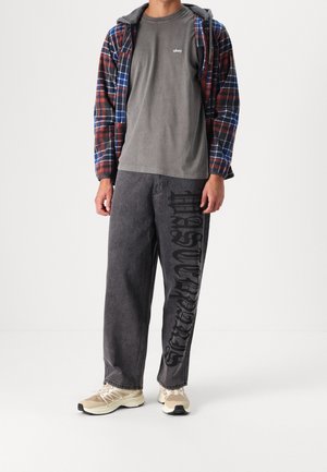 Punaisen, sinisen ja mustan ruudullinen flanellinen ylikangas; harmaa puuvillainen t-paita; mustat farkut, joissa suuret tekstit; vaaleat tennarit.