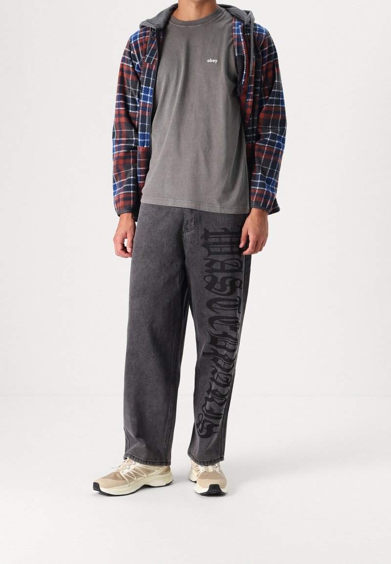 Chemise surchemise en flanelle à carreaux rouges, bleus et noirs ; t-shirt en coton gris ; jean noir avec grand texte ; baskets de couleur claire.