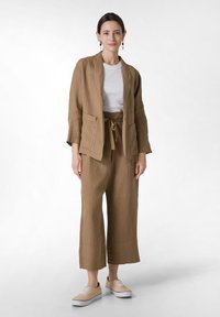 Ruskea pellavablazeri, jossa on kaksi etutaskua, yhdistettynä samaan sävyyn pellavahousujen kanssa. Valkoinen T-paita alla ja vaaleat kangaskengät.