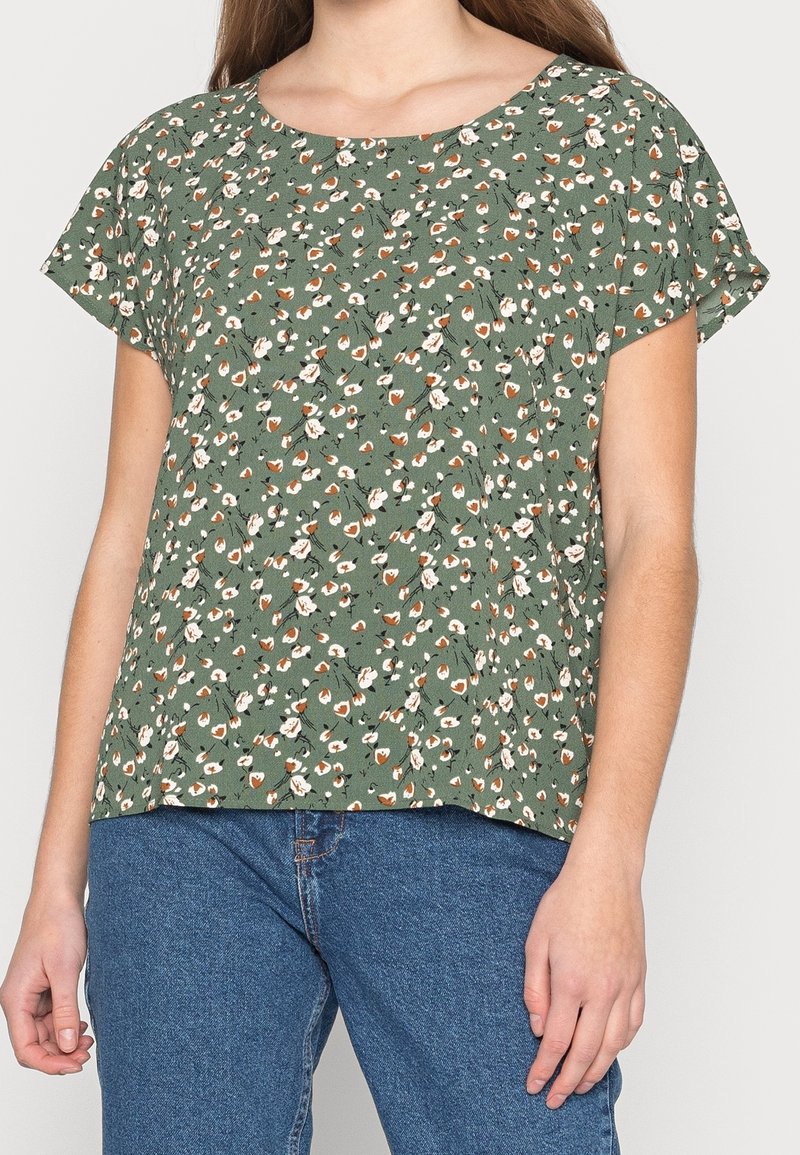 Kvinde iført en afslappet grøn bluse med hvidt og orange blomsterprint og blå jeans, stående foran en ensartet baggrund.