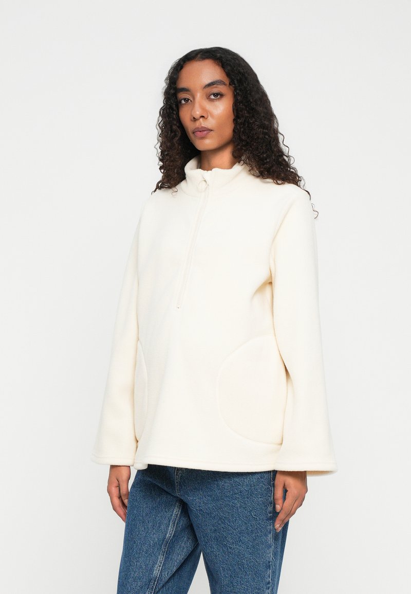 Donna con capelli ricci che indossa una giacca in pile color crema con zip frontale e jeans blu, in piedi su uno sfondo bianco semplice.
