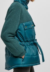 Tyrkysová péřová bunda s fleecovým vrškem, šňůrkovým pasem a kapsou na cvočky na hrudi. Má hladký, lesklý povrch a texturované rukávy.