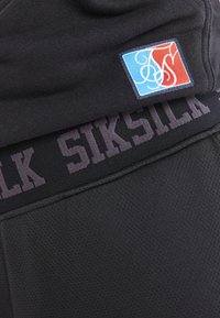 Nærbillede af sort sportstøjsstof med "SIKSILK" i taljen og en lille blå og rød lap med hvidt abstrakt design.