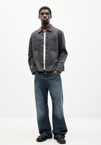 Graues Hemd mit Knöpfen und zwei Brusttaschen, darunter ein weißes T-Shirt, kombiniert mit hellblauen weit geschnittenen Jeans und schwarzen Schuhen.