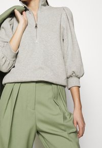 Sudadera de color gris claro con cuello de media cremallera y mangas abullonadas, combinada con pantalones plisados de color verde claro.