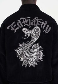 Musta corduroy-takki, jossa on broo'derattu käärme, joka on kietoutunut ruusuille, ja "Ed Hardy" -teksti yläpuolella hopealangalla.