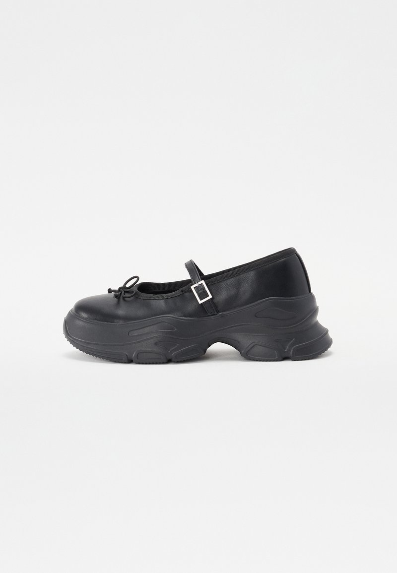 Black leather Mary Jane shoe with a rounded toe, bow detail, and an adjustable ankle strap. Features a chunky rubber sole with curved accents.