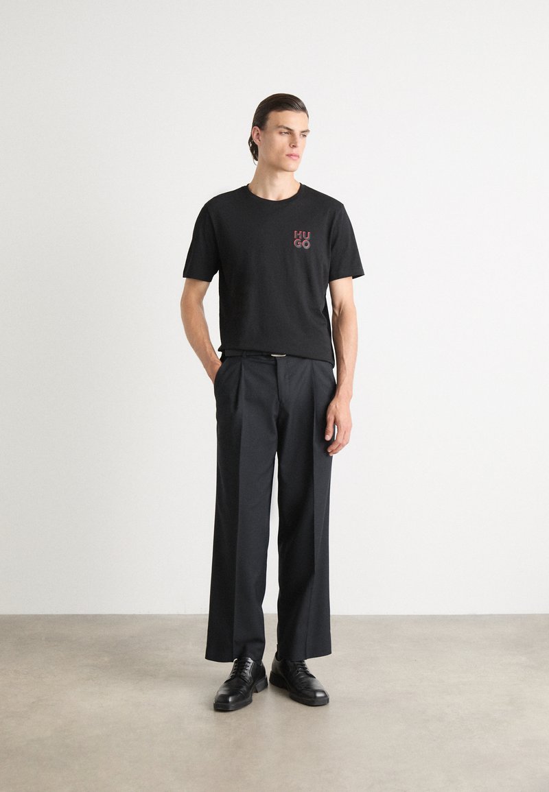 Zwarte katoenen T-shirt met een rode "HUGO" logo op de borst, gecombineerd met hoge zwarte broek en zwarte leren schoenen.