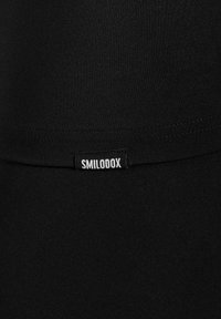 Tissu noir avec une petite étiquette noire cousue sur la couture, affichant le texte blanc "SMILODOX" en lettres majuscules.