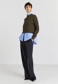 Maglione corto a treccia di colore verde oliva indossato sopra una camicia button-up blu chiaro, abbinato a pantaloni neri a gamba larga su misura e scarpe nere.