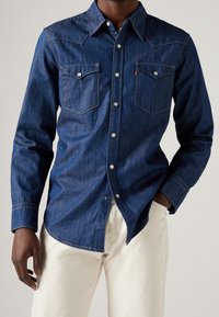 Mörk denimskjorta med långa ärmar, spetsig krage, två bröstfickor, silverfärgade tryckknappar och kontrasterande sömnadsdetaljer.