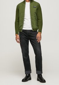 Grön vadderad zip-up jacka med ribbade manschetter över en vit skjorta, kombinerad med mörka jeans och svarta ankelskor. Anmärkningsvärd logotypdetalj på jackan.