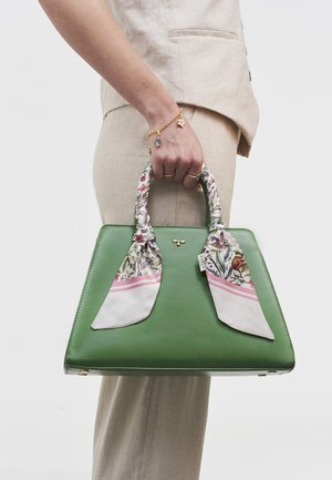 Roka tur zaļu somu ar ziedu raksta šalles rokturiem, valkājot bēša krāsas tērpu un zelta piekariņu rokassprādzi ar diviem gredzeniem.