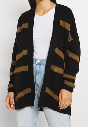 Sort cardigan med brune vandrette striber, oversized pasform, åben front, lange ærmer, lavet af blødt strikket materiale, båret over en hvid top.