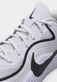 Λευκό αθλητικό παπούτσι Nike με μαύρο λογότυπο Swoosh, υφή στο πάνω μέρος, επενδεδυμένη γλώσσα και λευκά κορδόνια δεμένα σε σταυρωτό μοτίβο.