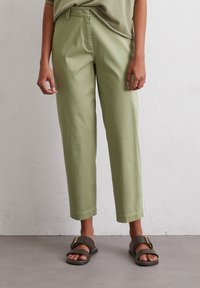 Light green cotton trousers with a relaxed fit, cropped length, and smooth texture. Paired with dark brown sandals featuring a gold buckle.