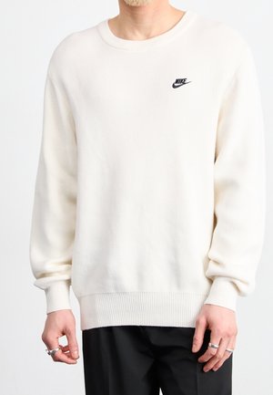Pull Nike blanc à manches longues porté par une personne avec des bagues aux deux mains, associé à un pantalon noir sur fond blanc.