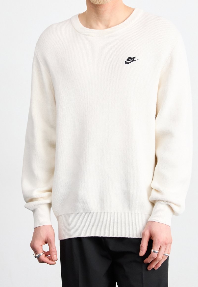 Pull Nike blanc à manches longues porté par une personne avec des bagues aux deux mains, associé à un pantalon noir sur fond blanc.