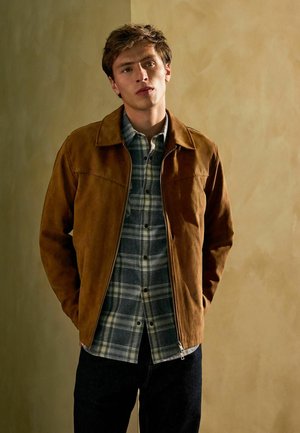 Veste en daim marron avec un col, portée par-dessus une chemise à carreaux bleue et verte. La veste présente une fermeture éclair et une coupe ajustée.
