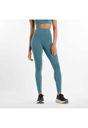 Femme portant un legging taille haute bleu sarcelle et un soutien-gorge de sport assorti, debout les mains détendues, chaussures de course noires, fond blanc.