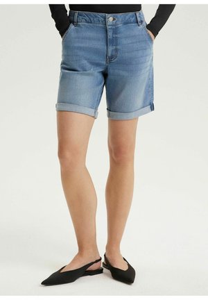 Jasnoniebieskie szorty jeansowe z podwiniętymi mankietami, noszone z czarnymi balerinami typu slingback i czarną bluzką z długim rękawem, ręce w kieszeniach, stojąca.