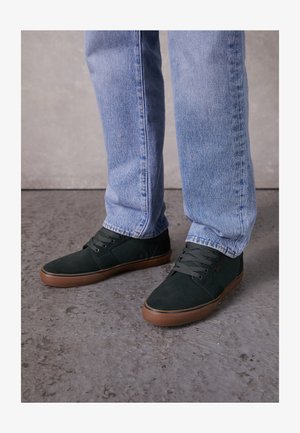 Baskets en suede vert avec semelles en caoutchouc marron, dotées d'un lacet et d'un col rembourré, associées à un jean en denim bleu clair.