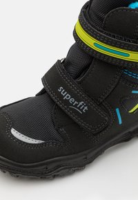 Bottine noire avec une finition texturée et des accents jaunes. Équipée d'une sangle en Velcro portant l'inscription "superfit" et d'éléments de design colorés.