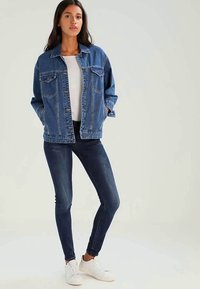 Τζην μπουφάν σε σκούρο μπλε χρώμα με δύο μπροστινές τσέπες και κλείσιμο με κουμπιά, φορεμένο πάνω από ένα λευκό μπλουζάκι και σε συνδυασμό με σκούρα στενά τζιν και λευκά αθλητικά παπούτσια.