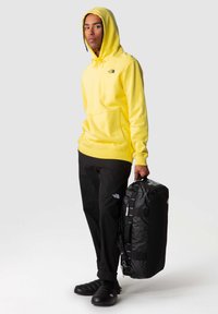 Yellow hooded sweatshirt with front pocket and logo; black pants; carrying black duffle bag with straps; black slip-on shoes.