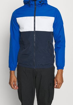 Mann trägt blaue, weiße und marineblaue Kapuzenjacke mit Reißverschluss und zwei Reißverschlusstaschen sowie schwarze Cargohosen vor neutralem Hintergrund.