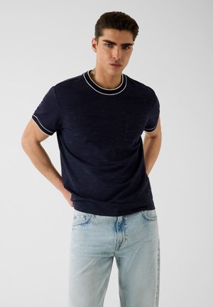 Jeune homme aux cheveux foncés portant un t-shirt bleu marine à bordure blanche et un jean bleu clair, debout les mains derrière le dos.