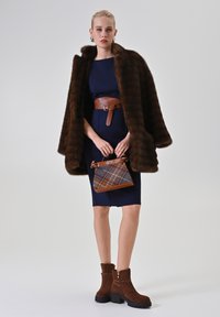 Brauner Kunstpelz-Cape mit einem marineblauen Kleid, das einen braunen Gürtel hat, braune Wildleder-Stiefeletten und eine karierte Handtasche mit Lederbesatz.