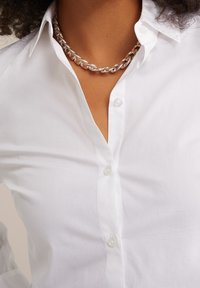 Vit skjorta med knappar och klassisk krage, med slät yta, klara knappar och ett silverfärgat kedjehalsband som accent.