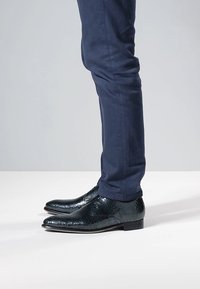 Kiiltävät mustat nahkakengät teksturoidulla pinnalla, kärjessä terävä ja matala korko, yhdessä laivastonsinisten housujen kanssa. Sivunäkymä.