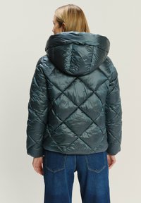 Prešívaná puffer bunda v tmavom tyrkysovom odtieni, s veľkou kapucňou a vertikálnymi švami. Lesklá, štruktúrovaná tkanina zdôrazňuje jej diamantový vzor.