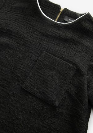 Top nero texturizzato con scollatura tonda, zip sul retro e tasca a toppa sul davanti. La superficie presenta un motivo ondulato.