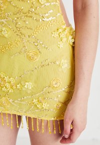 Gelbes Kleid mit floralem Stickmuster, Pailletten und Quastendetails, getragen von einer Person mit sichtbarem Arm und Hand.
