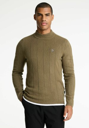 Mann in olivgrünem, geripptem Pullover und schwarzen Hosen, steht mit einer Hand in der Tasche vor einem einfarbigen Hintergrund.